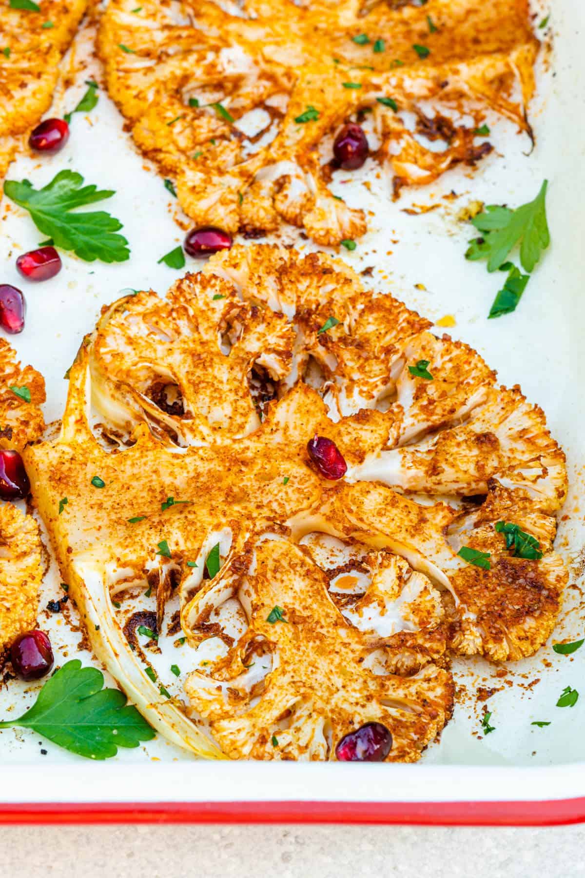 Close up of cauliflower steaks in an oven tray topped with pomegranate and parsley