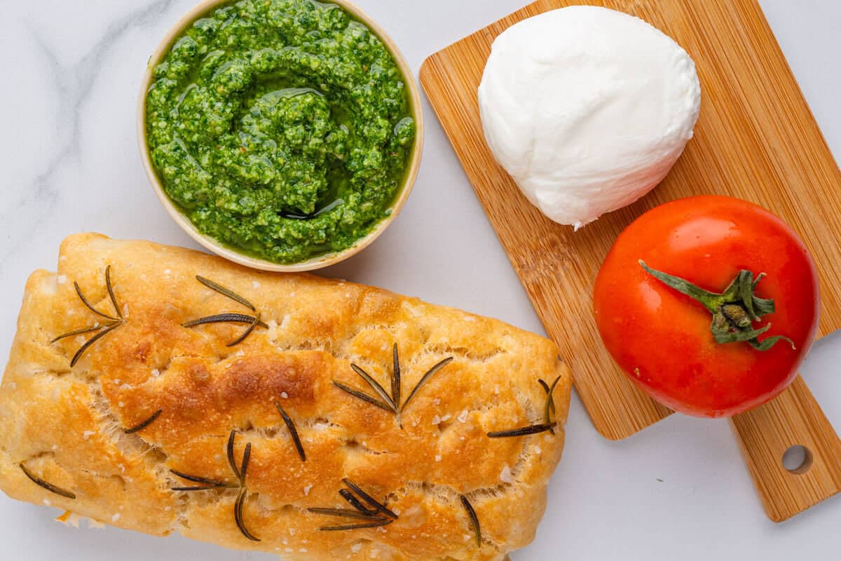 Ingredients to make a caprese sandwich including pesto, focaccia, mozzarella ball and tomato