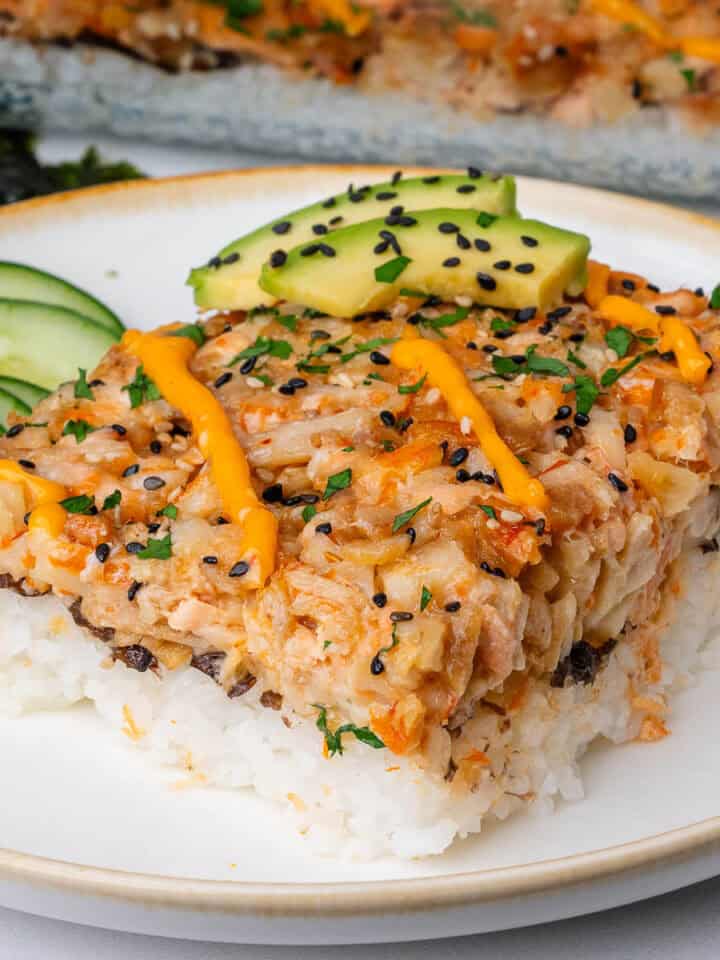 Piece of sushi bake on a plate topped with avocado, spicy mayo and sesame seeds