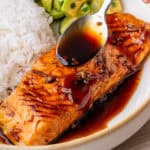 Honey garlic salmon with spoon of sauce being drizzled over the top and rice and salad on the side