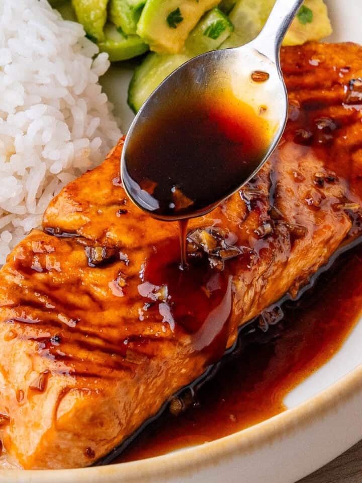 Honey garlic salmon with spoon of sauce being drizzled over the top and rice and salad on the side